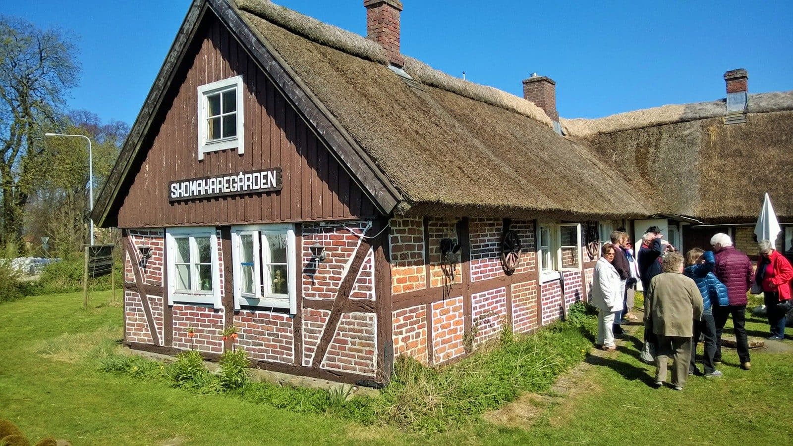 Skomakaregården i Allerum, ett korsvirkeshus med rött tegel och halmtak, med besökare på gården.
